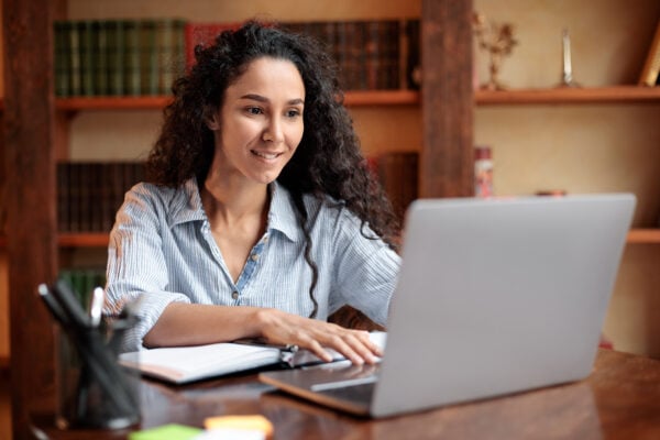 Doing Project. Portrait of curly young woman using laptop computer and typing on keyboard, writer sitting at table. Female manager working remotely or studying online at home, telecommuting